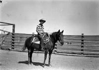 [Young cowboy on horseback in front of pole fence]