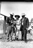 [Unidentified Cowgirl stands between two formally attired men one of whom is looking at photographs]