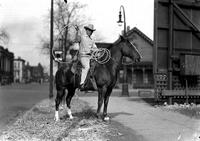 [Unidentified cowboy atop stationary horse and holding rope loop to side behind billboard]