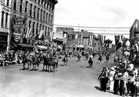 [Two cowgirls on horseback flanked by two military personnel on horseback parading down street]