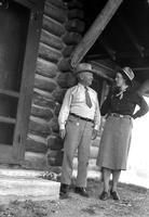 [Unidentified elderly man and woman outside log cabin]