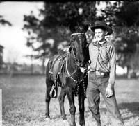 [Unidentified young man in flat-crowned hat standing with saddled horse trees in background]