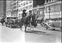 [Unidentified cowgirl & possibly Florence Randolph atop horses making way down busy city street]
