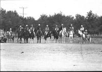 [Line of unidentified cowboys & one cowgirl on horseback with a child on a pony at each end]