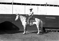 [Unidentified Cowboy with hands on saddle horn atop horse; wooden plank fence behind him]