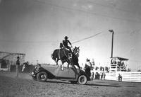 [Unidentified Cowboy doing Roman Stand jump over auto as Clown sits on spare tire housing]