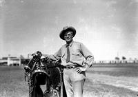 [Unidentified Cowboy leaning on silver mounted saddle in grassy field, stadium in background]