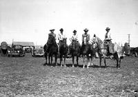 [Group of five unidentified cowboys on horseback, autos and trailers in background]