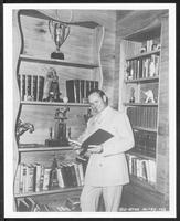 Gene Autry in his favorite room of his San Fernando Valley home (den), with trophies and cups he has won as America's favorite singing cowboy. He looks through album of photos from his Columbia release "On Top of Old Smoky."