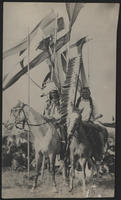 [2 Native Americans on horseback in full dress,one on right is holding a lance with feathers on it]