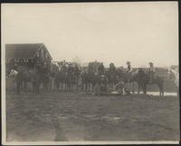 [2 Cowboys heading and heeling with 20 other cowboys, all near a barn]