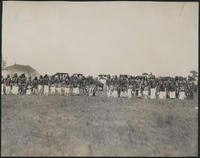 [26 Native American men in full dress with whistles in their mouths, spectators and wagons behind]