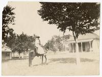 Cowgirl on sidesaddle horseback under tree near house]