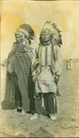 [Two Southern Ute men in headdresses, blankets, jewelry, knee bells, and traditional clothing]