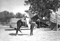 [Unidentified older cowboy wearing holster on horse posed in front of trucks & trailers]