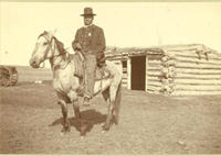 [Indian policeman posed on horse in front of log building]