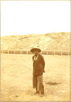 [Standing elderly Indian man wearing necklace with fence and rocky hill behind]