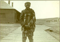 [Single elderly Indian man standing on boardwalk with buildings and street behind]