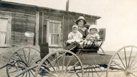 [Unidentified woman sitting with two toddlers in carriage in front of home]