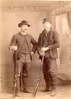 [Two posed hunters standing with rifle and shotgun while smoking]