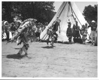 [Pair of male Indian dancers with singers/drummers & tipi in background]