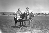 [Unidentified cowgirl and cowboy posed on horses with ranch buildings behind]