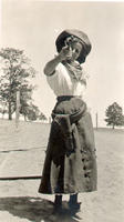 [Unidentified woman dressed as a cowgirl wearing skirt, holster, and hat with gun drawn]
