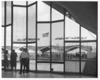 [View from museum interior through windows to fountains & flags]