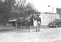 [Unidentified cowboy posed by saddled horse in dirt road by automobile]