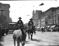 [Two unidentified cowboys on horses on a street lined with buildings]