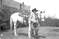[Unidentified older cowboy holding coiled rope & standing by saddled horse]