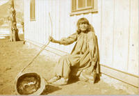 [Sitting Indian woman with burden basket on ground in front of building]
