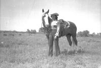 [Unidentified cowgirl posed and standing beside saddled horse]