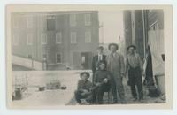 [5 men sitting and standing in front of a tent near a building]