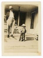 [Man with little boy, (boy is dressed as a cowboy), standing on a front porch]