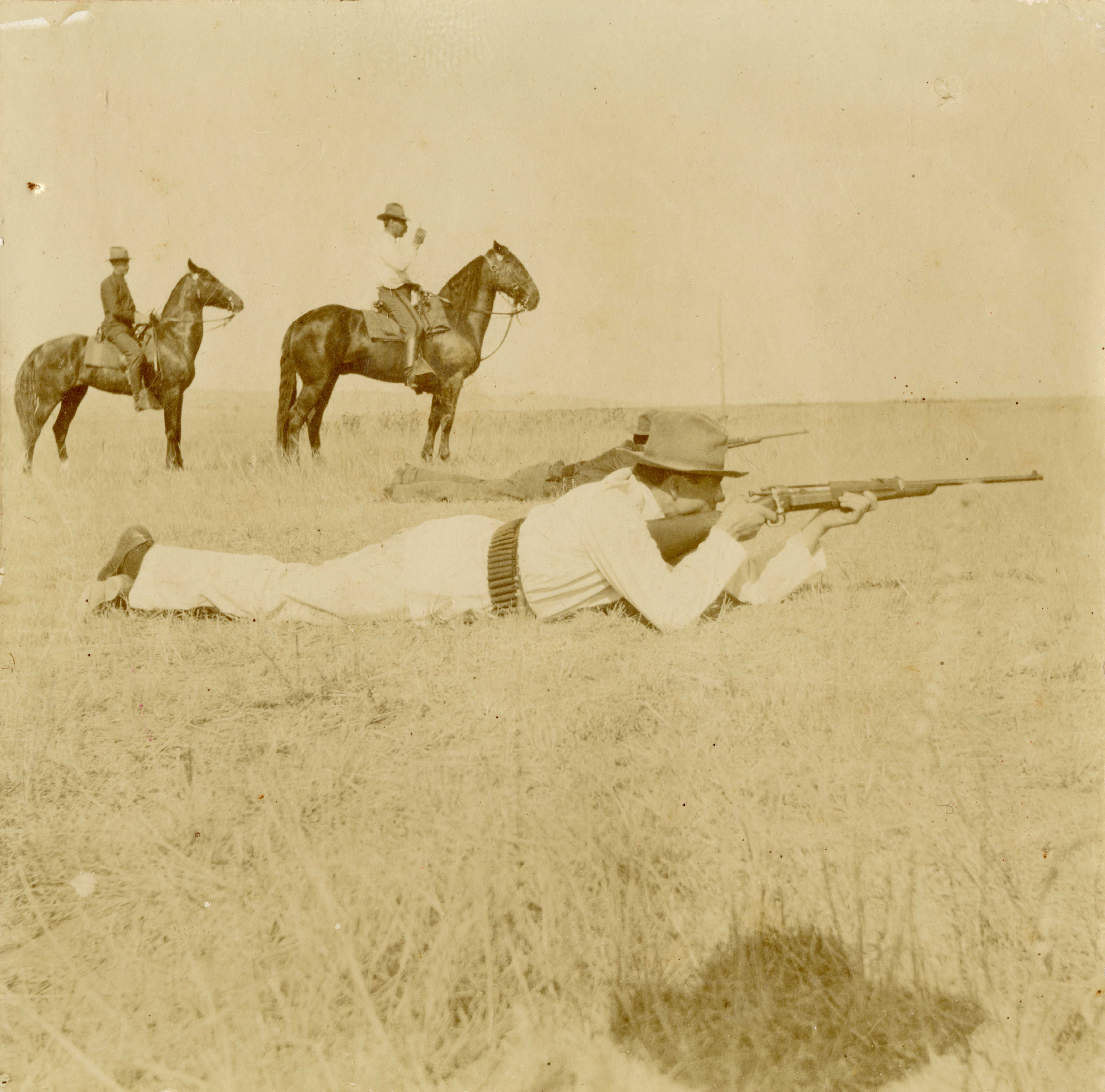 [Soldier shooting in the prone position, two men  on horseback behind him]