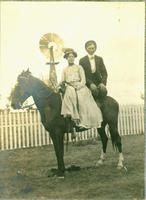 [Well-dressed man and woman sharing the back of a horse with fence and windmill in background]