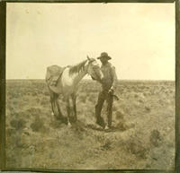 [Cowboy standing beside white horse which is blindfolded and has blanket on back]
