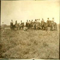 [1 Squatting Cowboy in front of a line of 13 cowboys on horseback]