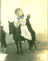 [Well dressed man and woman sharing the back of a horse in front of fence and windmill]