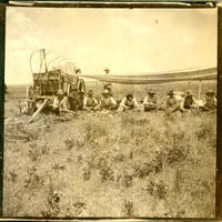 [9 sitting cowboys sitting and eating beneath tarp extended from chuck wagon]