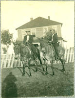 [Two well-dressed cowboys posing on horseback in front of house and fence]