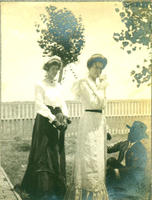 [Two well-dressed women standing near sitting well-dressed man]
